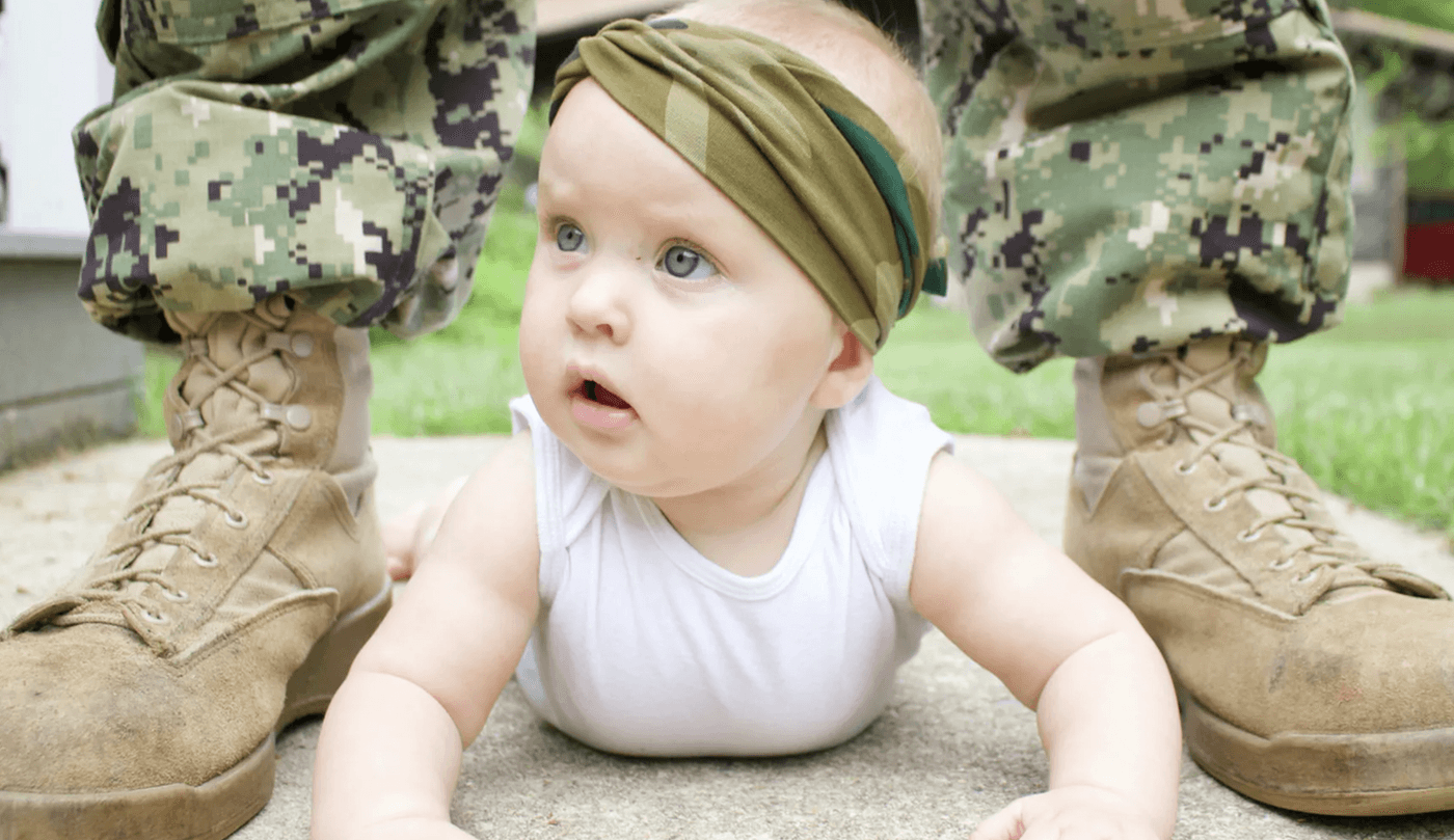 Infant with a military head wrap crawling on the ground in between the boots and legs of a military serviceman.